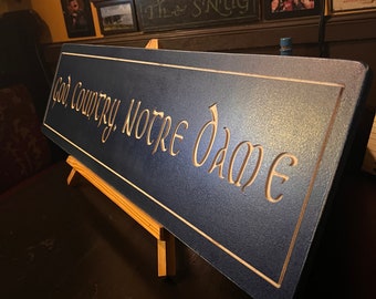 May include: A dark blue wooden sign with the words "God Country, Notre Dame" carved in gold lettering. The sign is on a small wooden easel.