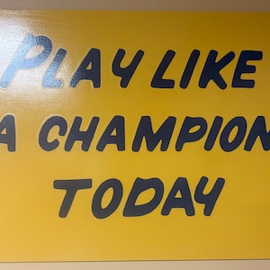 May include: A yellow sign with the words "PLAY LIKE A CHAMPION TODAY" in dark blue lettering. The sign has a glossy finish and is rectangular in shape. The text is in a bold, sans-serif font.