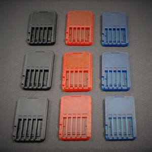 May include: Nine plastic cases in a grid arrangement. Three are black, three are red-orange, and three are blue. Each case has a rectangular shape with a series of vertical slots on one side. The cases are likely for storing small items.