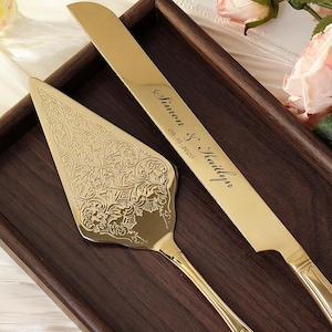 Puede incluir: Un juego de cuchillo y servidor de pastel dorado con detalles florales ornamentados. El cuchillo está grabado con "Simon & Kaitlyn" y la fecha "09.19.2026". El juego se exhibe en una bandeja de madera oscura, ideal para bodas.