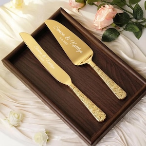 May include: A gold-toned cake knife and server set with ornate floral handles. The blades are engraved with the text Simon & Kaitlyn and the date 9-6-2025. The utensils are placed inside a dark brown wooden rectangular tray.