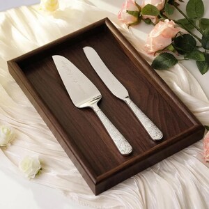 May include: A silver cake server and a matching serrated cake knife with ornate, textured handles. Both pieces feature the engraved text "Simon & Kaitlyn" on the blades. The utensils are positioned on a rectangular dark brown wooden tray.