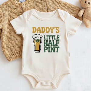 May include: Cream-colored baby bodysuit with the text "Daddy's Little Half Pint" in gold and green. A beer glass graphic is included. A tan knitted sweater and a small teddy bear are in the background.