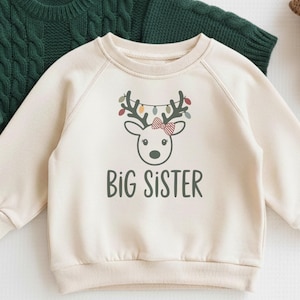 May include: A cream-colored sweatshirt with the text "BIG SISTER" and a reindeer graphic with a red bow and string lights. A green cable-knit sweater is behind it. A small crocheted reindeer is to the right.