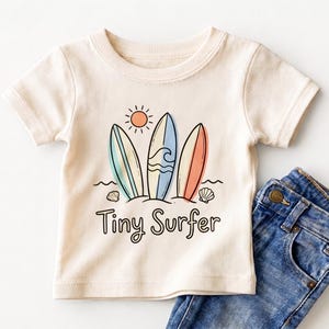 May include: A cream-colored short-sleeve t-shirt featuring a graphic of three surfboards, a sun, and seashells above the text Tiny Surfer. The shirt is paired with blue denim jeans showing a metal button and a pocket seam detail.