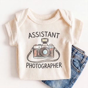 May include: Cream-colored baby onesie with the words "ASSISTANT PHOTOGRAPHER" above and below a colorful illustration of a camera. The onesie is paired with a pair of blue denim jeans.