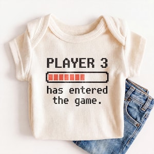 May include: Cream-colored baby onesie with the text "PLAYER 3 has entered the game." The text is in a pixelated font, with a red loading bar graphic. Blue denim jeans are partially visible.