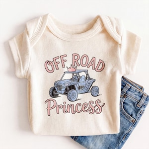 May include: Cream-colored baby onesie with the words "OFF ROAD Princess" in pink script and a blue off-road vehicle graphic. The onesie is paired with a pair of blue denim jeans.
