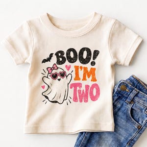 May include: A cream-colored short-sleeve t-shirt featuring a graphic of a ghost wearing heart-shaped sunglasses and a pink bow. The text reads BOO! I'M TWO in black, orange, and pink distressed lettering, accompanied by a small black bat and hearts.