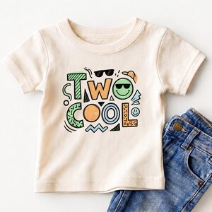 May include: A cream-colored toddler t-shirt with the words "TWO COOL" in a colorful, playful design. The graphic includes sunglasses, a smiley face, and other fun shapes. Blue denim jeans are visible on the right.