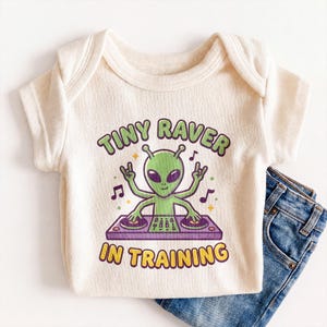 May include: A cream-colored ribbed short-sleeve bodysuit featuring a graphic of a green four-armed alien DJing on a purple turntable. The text reads "TINY RAVER IN TRAINING" in green and yellow letters, accompanied by blue denim jeans.
