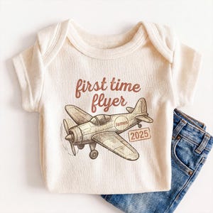 May include: A cream-colored ribbed short-sleeve bodysuit featuring a brown graphic of a vintage airplane. The text reads "first time flyer" above the plane, with the name "James" inside a circle on the fuselage and the year "2025" in a box.