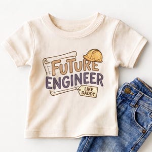 May include: A cream-colored short-sleeve t-shirt featuring a graphic of a blueprint, a yellow hard hat, and the text "FUTURE ENGINEER" with a small tag that reads "LIKE DADDY" next to a pair of blue denim jeans with a visible brass button.