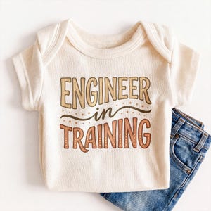 May include: A cream-colored ribbed short-sleeve bodysuit featuring the text "ENGINEER in TRAINING" in brown and tan gradient lettering with a decorative curved line and small dots, paired with a piece of blue denim fabric at the bottom right.