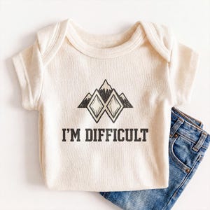 May include: Cream-colored baby onesie with the text "I'M DIFFICULT" in black block letters. The onesie features a graphic of three mountain peaks and two diamond shapes. A pair of blue denim jeans is partially visible.