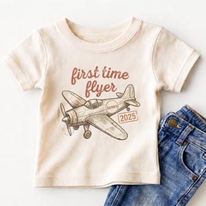May include: A cream-colored short-sleeve t-shirt featuring a brown sketch of a propeller airplane. The text "first time flyer" is printed above the plane, with the name "James" inside a circle on the fuselage and "2025" in a rectangular box.