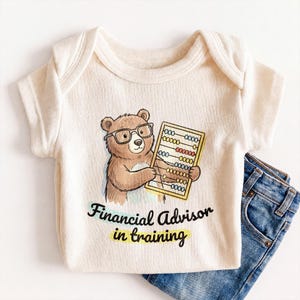 May include: A cream-colored ribbed short-sleeve bodysuit featuring a graphic of a brown bear wearing glasses and holding an abacus. Below the bear, the text reads Financial Advisor in training in black letters with a yellow highlight underneath.