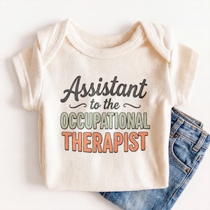 May include: Cream-colored baby onesie with the text "Assistant to the Occupational Therapist" in black, green, and orange. The onesie is paired with a folded pair of blue denim jeans.