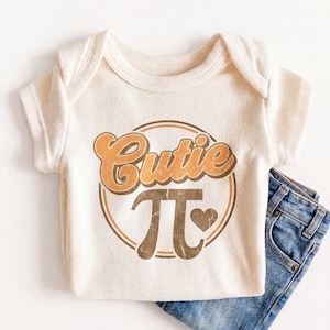 May include: A cream-colored ribbed short-sleeve bodysuit featuring a circular graphic with the text Cutie and the mathematical symbol pi in brown and orange tones, accompanied by a small brown heart icon, paired with blue denim jeans.