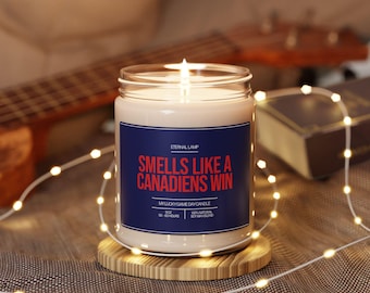May include: A lit candle in a glass jar with a blue label that reads "Smells Like a Canadiens Win." The candle sits on a wooden coaster, with string lights and a guitar in the background. The candle is 255 grams and made of a soy wax blend.