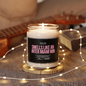 May include: A lit candle in a glass jar with a black label that reads "Smells Like An Inter Miami Win." The candle sits on a wooden coaster, surrounded by string lights. A guitar and a book are in the background.