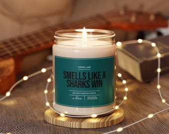 Pode incluir: Uma vela acesa num frasco de vidro com um rótulo verde-azulado que diz "Smells Like a Sharks Win". A vela está sobre um descanso de madeira, com uma guitarra e um livro no fundo. A vela é rodeada por uma cadeia de luzes.