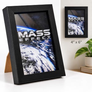 May include: A black rectangular shadow box frame containing a 4 inch by 6 inch print of the Mass Effect logo over a view of Earth from space. The frame has a thick black border and a light brown easel back support for standing on a flat surface.