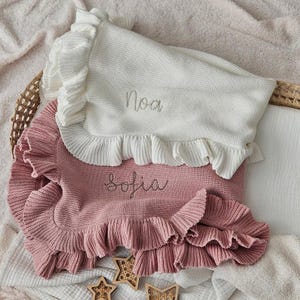 May include: Two knitted baby blankets, one white and one pink, with ruffled edges. The white blanket has the name "Noa" embroidered in gray, and the pink blanket has "Sofia" embroidered. The blankets are folded and placed in a woven basket.