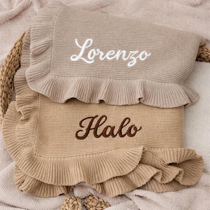May include: Two beige knitted baby blankets with ruffled edges. One blanket has the name "Lorenzo" embroidered in white, and the other has "Halo" embroidered in brown. The blankets are folded and stacked.