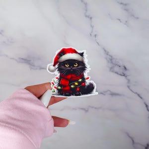 May include: A sticker featuring a black cat wearing a Santa hat and a red scarf, adorned with Christmas lights. The cat has yellow eyes and a fluffy appearance, perfect for holiday decor.