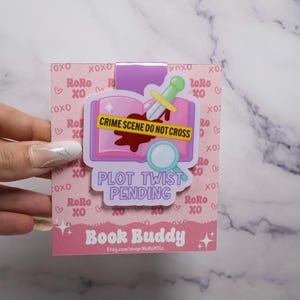 May include: A pink and purple Book Buddy bookmark with a crime scene design. The bookmark features an open book, a knife, and a magnifying glass, with the text "CRIME SCENE DO NOT CROSS" and "PLOT TWIST PENDING".