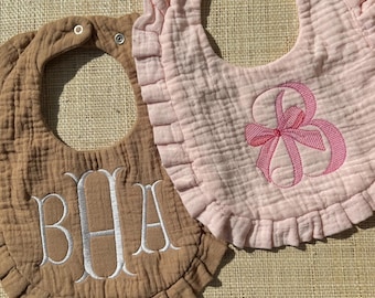 May include: Two muslin baby bibs with ruffled edges and snap closures. The tan bib features white embroidered monogram letters BHA in a script font. The light pink bib features a pink embroidered bow design centered on the front fabric.