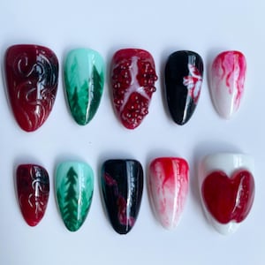 May include: A collection of ten artificial nails with diverse designs. The nails showcase colours like red, black, white, and teal. Designs include a pomegranate, a forest, and abstract patterns. The nails are oval-shaped.