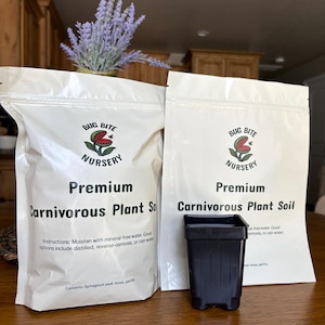 May include: Two white bags of "Premium Carnivorous Plant Soil" from Bug Bite Nursery, with instructions and contents listed. A black square plant pot sits in front of the bags. A vase of purple flowers is in the background.