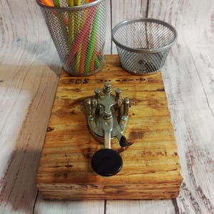Puede incluir: Una llave telegráfica vintage montada sobre una base de madera, con las letras "SOS" visibles. También hay dos portalápices de malla metálica y una colección de lápices de colores. La llave es plateada con un botón negro.