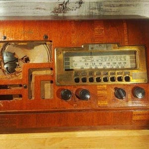 Puede incluir: Una radio de madera vintage con acabado marrón oscuro, que presenta un panel de dial rectangular en tono dorado con texto negro y marcas de frecuencia. El frente incluye cuatro perillas de control negras y una rejilla de altavoz con tela rasgada.