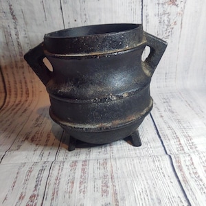 May include: A black cast iron cauldron with two handles and three small legs. The cauldron has a round body and a wide opening. The exterior has a textured, aged appearance, suggesting it is vintage or antique.