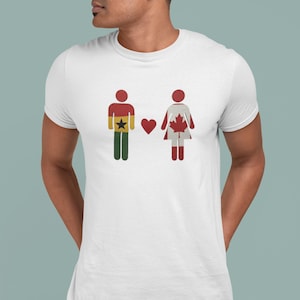 Könnte beinhalten: Weißes T-Shirt mit einer Grafik von zwei Strichmännchen, die ein Paar darstellen. Eine Figur ist in den Farben der ghanaischen Flagge gestaltet, die andere in den Farben der kanadischen Flagge, mit einem roten Herzen dazwischen. Das T-Shirt wird von einer Person getragen.