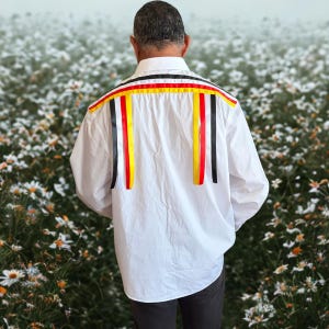 May include: A white long-sleeved button-down shirt featuring horizontal and vertical ribbon trim across the back shoulders. The ribbons are arranged in repeating stripes of black, white, red, and yellow, creating a decorative pattern.