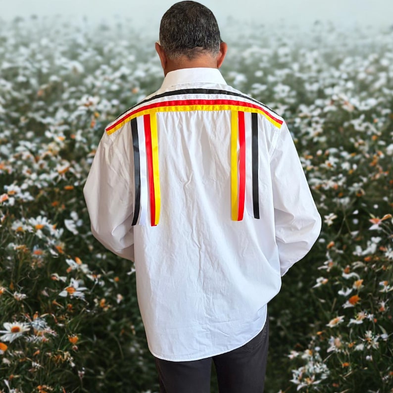 May include: A white long-sleeved button-down shirt featuring horizontal and vertical ribbon trim across the back shoulders. The ribbons are arranged in repeating stripes of black, white, red, and yellow, creating a decorative pattern.