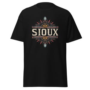 Puede incluir: Camiseta negra con la palabra "SIOUX" en una fuente desgastada. El texto está centrado y enmarcado por un diseño decorativo con acentos rojos, azules y amarillos, creando una estética de inspiración nativa americana.