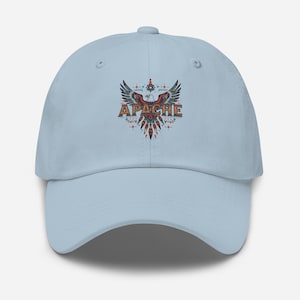 May include: A light blue baseball cap featuring a colorful graphic of an eagle with outstretched wings and the word "APACHE" in a stylized font. The design incorporates red, blue, and orange hues, creating a striking visual contrast against the cap's light background.