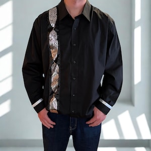 May include: A black long-sleeve button-down shirt featuring a vertical panel on the right side decorated with a repeating silver metallic feather pattern. The shirt includes a standard collar, a front button placket, and silver bands on the cuffs.