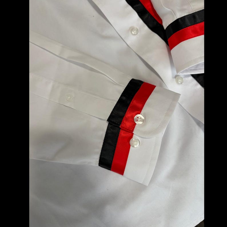 May include: A crisp white long-sleeve button-down shirt featuring a distinctive decorative band on the cuffs. The cuffs are accented with a bold horizontal stripe pattern consisting of a black satin ribbon followed by a vibrant red satin ribbon.