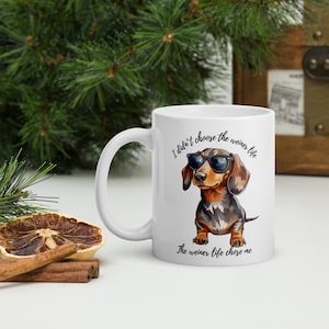 Può includere: Tazza in ceramica bianca con un disegno di un bassotto con occhiali da sole. La tazza ha il testo "I didn't choose the weiner life, the weiner life chose me". Il cane è marrone, nero e marrone chiaro.