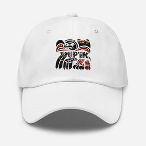 Op de afbeelding: Een witte baseballcap met een zwart, rood en wit grafisch ontwerp met het woord "YUP'IK". Het ontwerp bevat een gestileerde vogel en andere traditionele elementen. De cap heeft een gebogen klep.