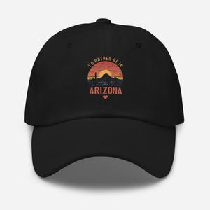 May include: Black baseball cap with a graphic design featuring a sunset over mountains and the text "I'd rather be in Arizona." The design includes a heart symbol. The cap is made of a durable fabric and has a curved brim.