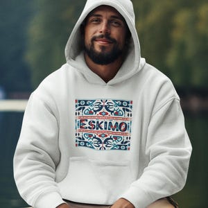 May include: White hooded sweatshirt with a colorful design featuring the word "ESKIMO" in the center. The design includes red, blue, and white patterns. The person is wearing a bracelet.