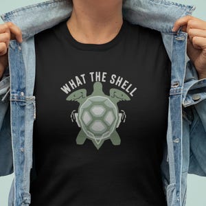 Könnte beinhalten: Schwarzes T-Shirt mit einem Schildkröten-Grafik und dem weißen Schriftzug "WHAT THE SHELL". Das Schildkröten-Design ist grün und grau. Die Person trägt eine Jeansjacke.