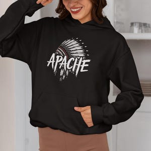 Puede incluir: Sudadera con capucha negra con un diseño de tocado nativo americano blanco y rojo y la palabra "APACHE" en blanco. La sudadera con capucha tiene un bolsillo delantero y una capucha con cordón. La persona de la imagen lleva una gorra de béisbol vaquera.
